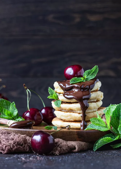 Stack of fluffy pancakes or fritters with chocolate and cherry jam, fresh cherry and mint. Delicious dessert for breakfast. Dark stone background, rustic style